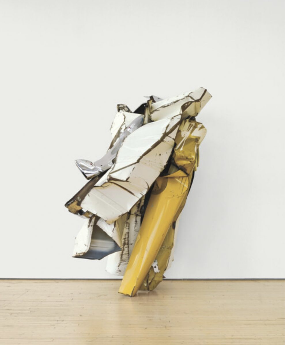 Color photograph of sculpture by John Chamberlain, called “The Hot Lady from Bristol,” circa 1979. White, silver, and yellow painted and chromium-plated steel plates are crushed together to create a sculptural form which leans upright against the white wall behind it. The edges are rusty, showing depth and folds in each steel plate.